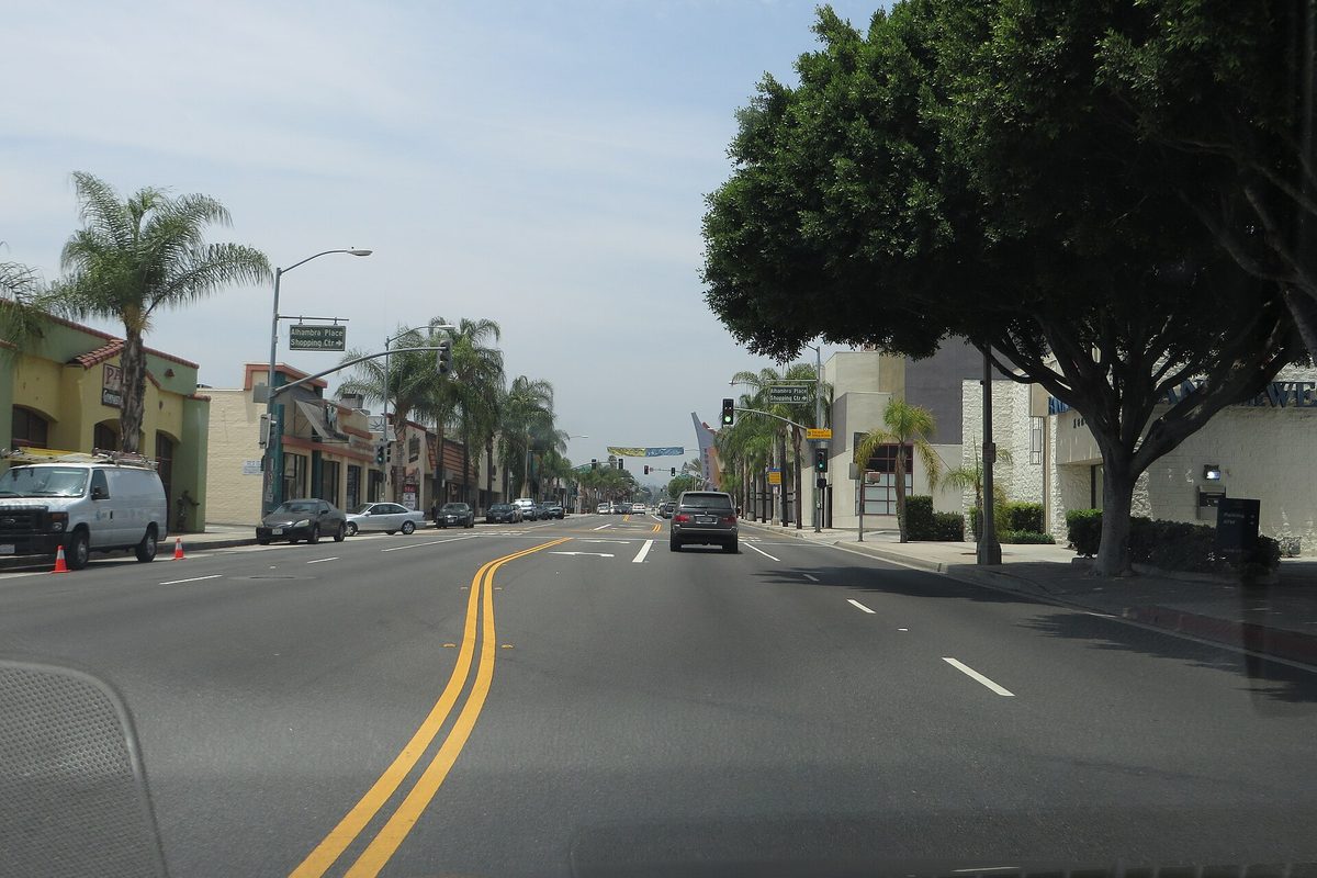 A wide four-lane avenue with a double yellow center line, palm trees and low storefronts under a hazy sky