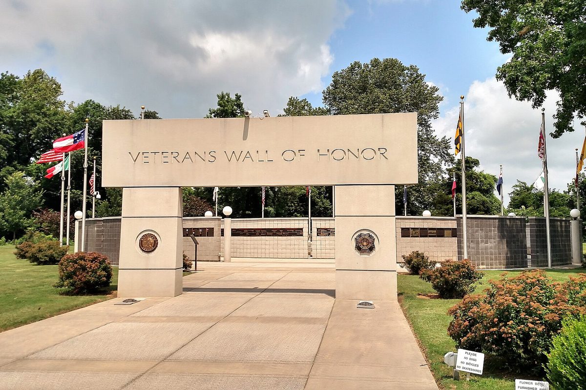 The stone Veterans Wall of Honor entrance arch in Bella Vista, with flags flying on both sides and shrubs along the walkway