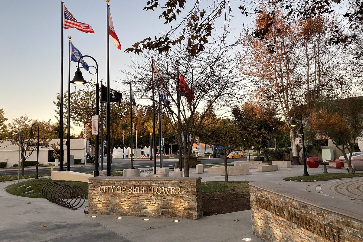 The Bellflower Veterans Memorial at Library Garden park at dusk, with a stone wall reading City of Bellflower, flagpoles and autumn trees