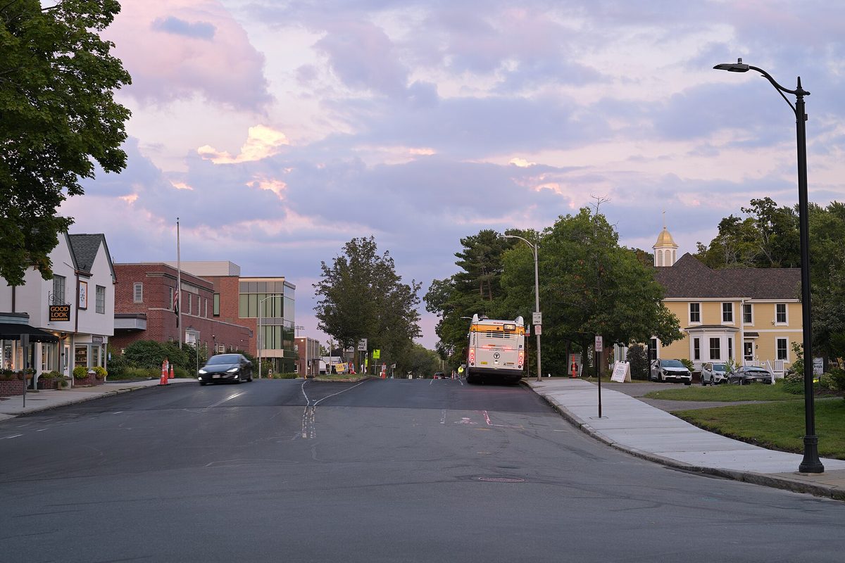 Concord Avenue in Belmont at dusk under pink clouds, with an MBTA bus at the curb and a yellow house with a cupola on the right