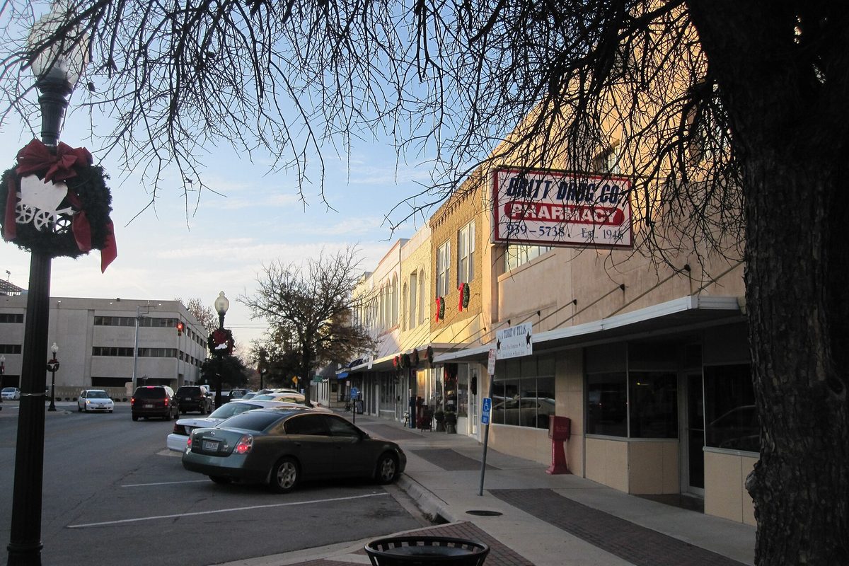 Downtown Belton storefronts at dusk, with a holiday wreath on a lamppost, an old Pharmacy sign over the sidewalk and cars parked in angled spaces