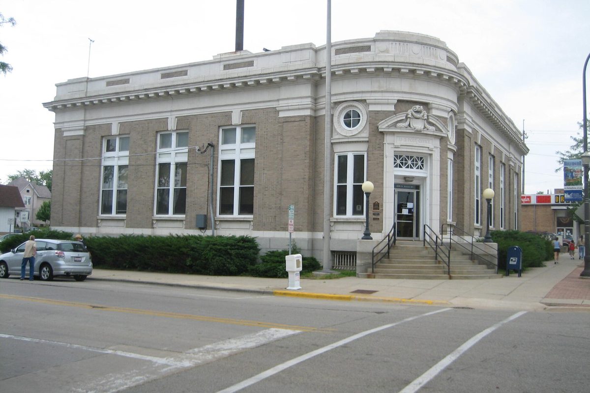 Belvidere's historic U.S. Post Office, a tan brick and stone corner building with a carved entry and lamp posts, a silver car parked at the curb