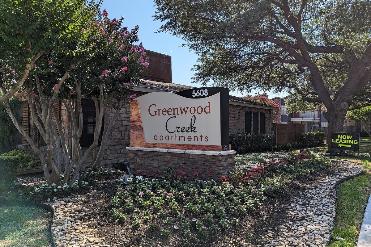 The Greenwood Creek Apartments entrance sign in Benbrook, set in a bed of pink and white flowers and river stones under a big oak and blooming crape myrtles