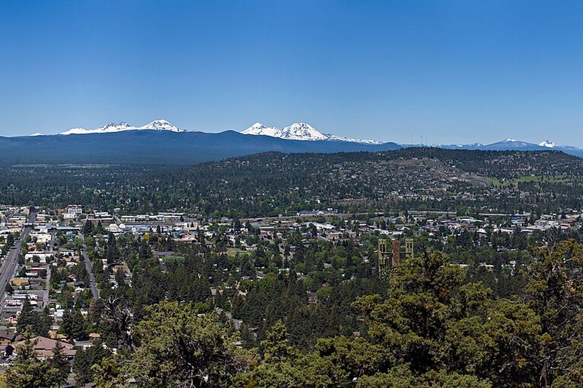 Bend spread across a pine forest below a row of snow-capped Cascade peaks under a clear blue sky, seen from Pilot Butte