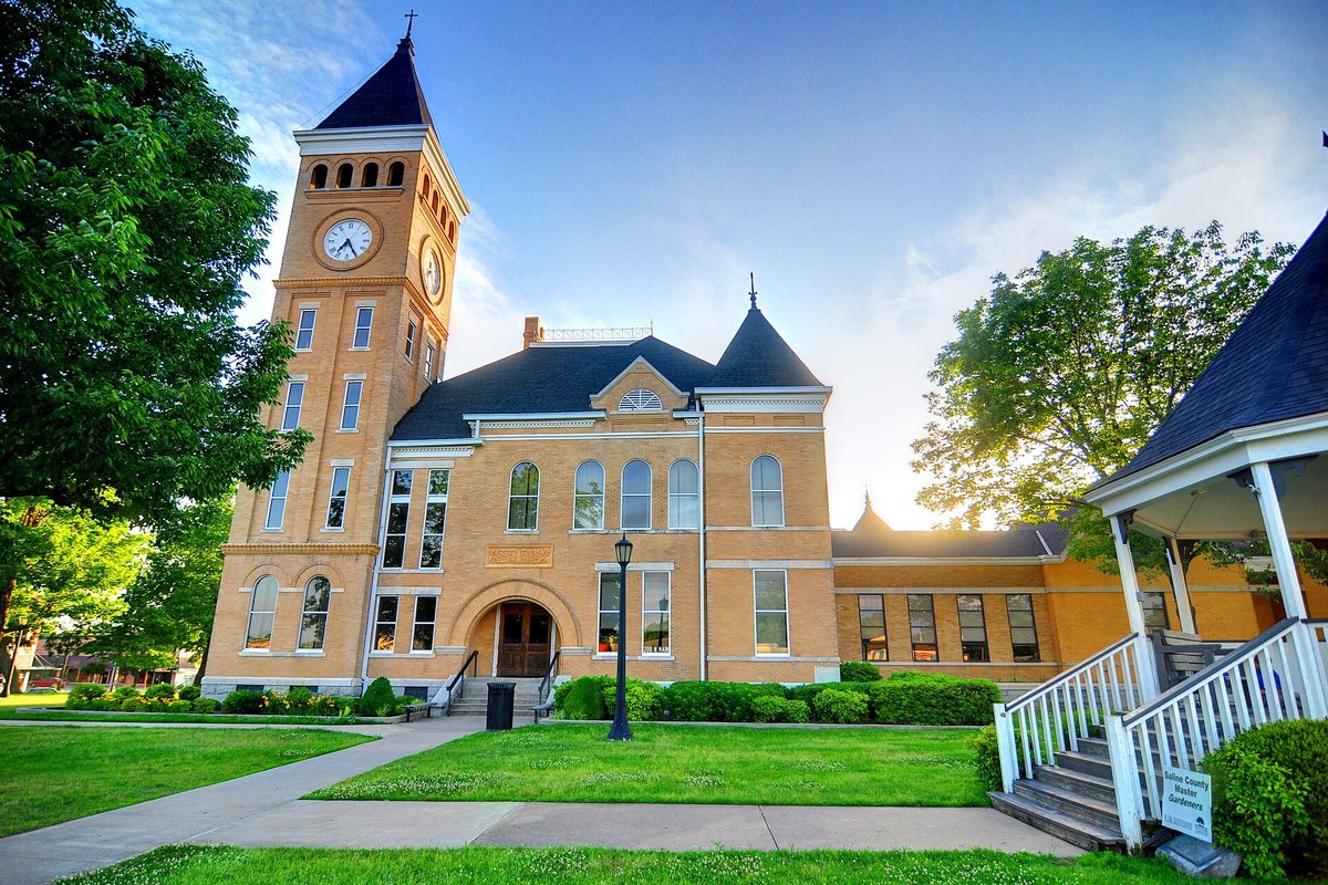 The yellow-brick Saline County Courthouse with its square clock tower and arched entrance, behind a green lawn under a blue sky