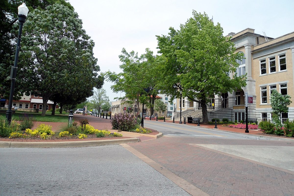 A brick-paved street on the downtown square in Bentonville, with flower beds, leafy trees, street lamps and a cream stone building with front steps