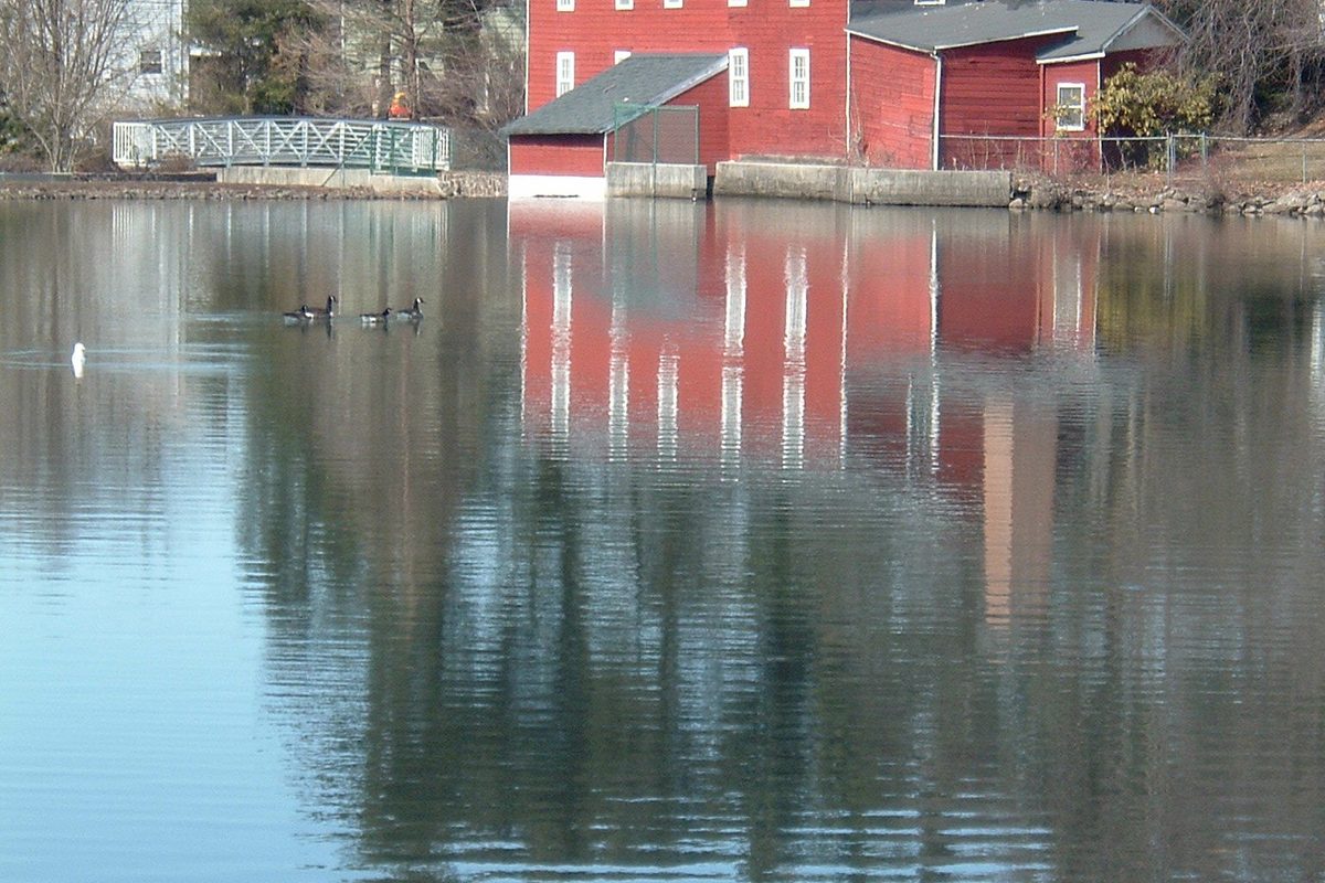 Cooper's Pond in Bergenfield, with a red barn-style building and its reflection on the still water and a pair of geese swimming on the left