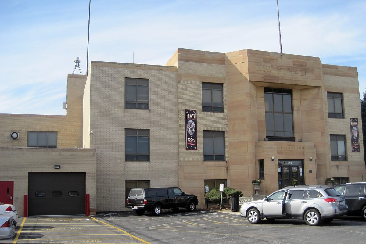 Berwyn's tan stone Municipal Building with City Hall carved above the entrance, a garage door on the left and cars in the parking lot in front