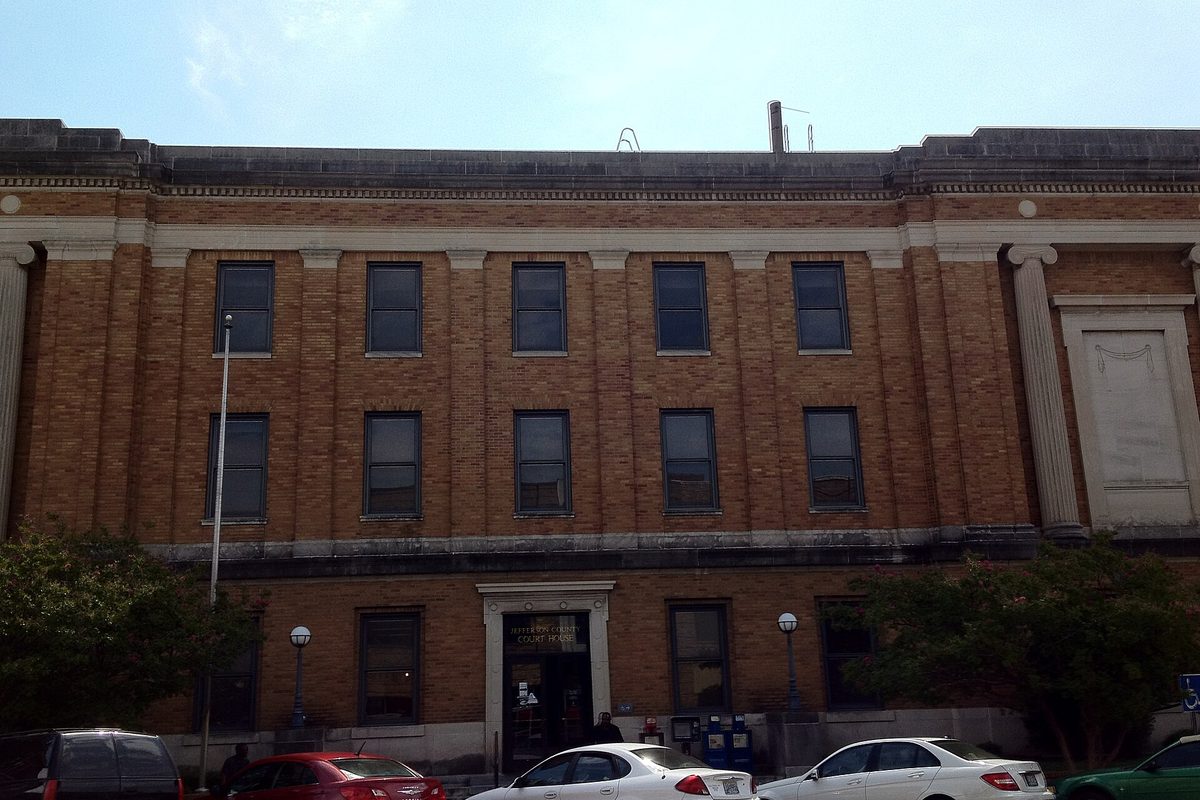 The tan brick, three-story Jefferson County Courthouse in Bessemer with stone pilasters, its front door shaded by trees and cars parked along the curb