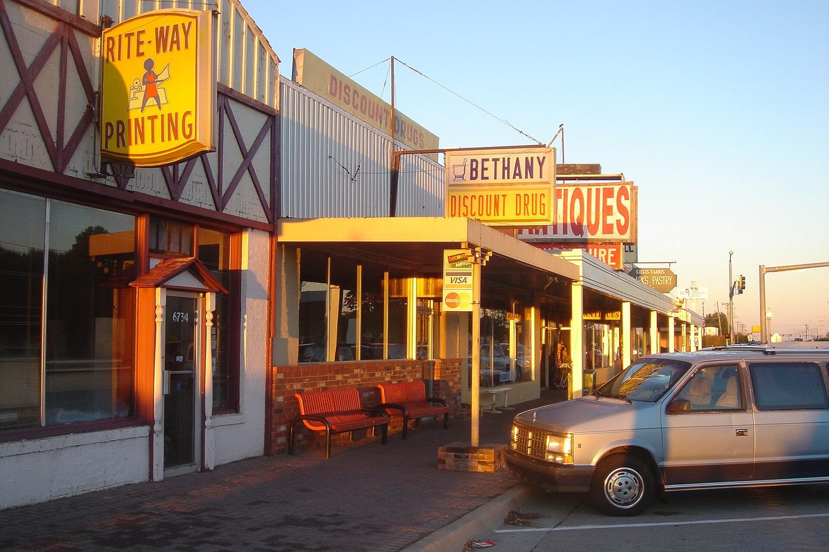 Storefronts on a Bethany street at sunset, with signs for Rite-Way Printing, Bethany Discount Drug and antiques, red benches under the awning and a minivan at the curb