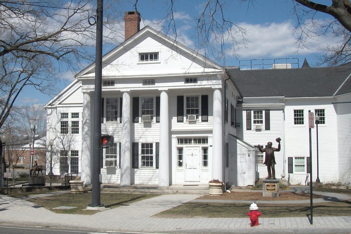 The white Greek Revival Seth Seelye House in Bethel, now a public library, with tall columns, a bronze statue of a man waving a hat on the lawn and a red fire hydrant by the sidewalk