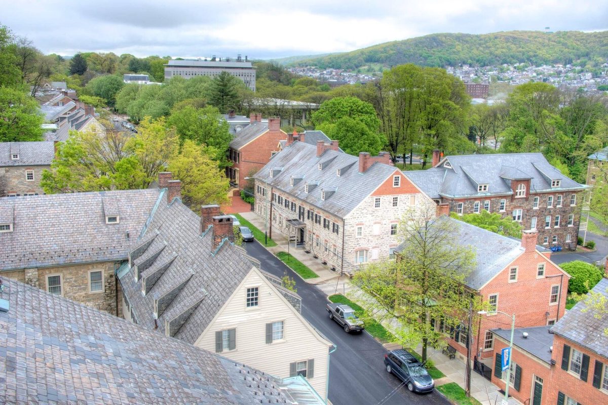 View from a church belltower over Bethlehem's historic stone and brick buildings with slate roofs, a tree-lined street with parked cars, and a wooded hill beyond