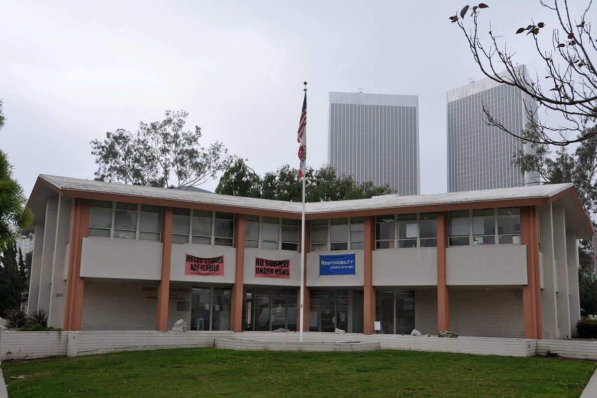 The two-story Beverly Hills Unified School District headquarters with salmon-colored pillars, a flagpole and a lawn, two tall towers behind it