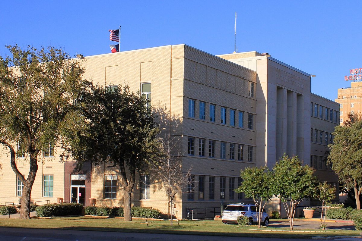 The tan, flat-roofed Howard County courthouse in Big Spring with tall vertical window bands, American and Texas flags on the roof, and trees and a white SUV in front