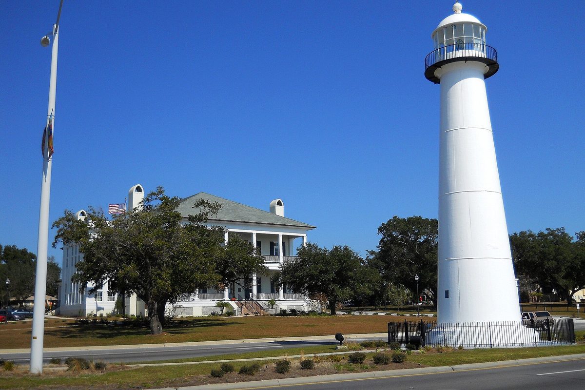 The white Biloxi Lighthouse beside U.S. Highway 90, with the white two-story visitors center behind live oaks