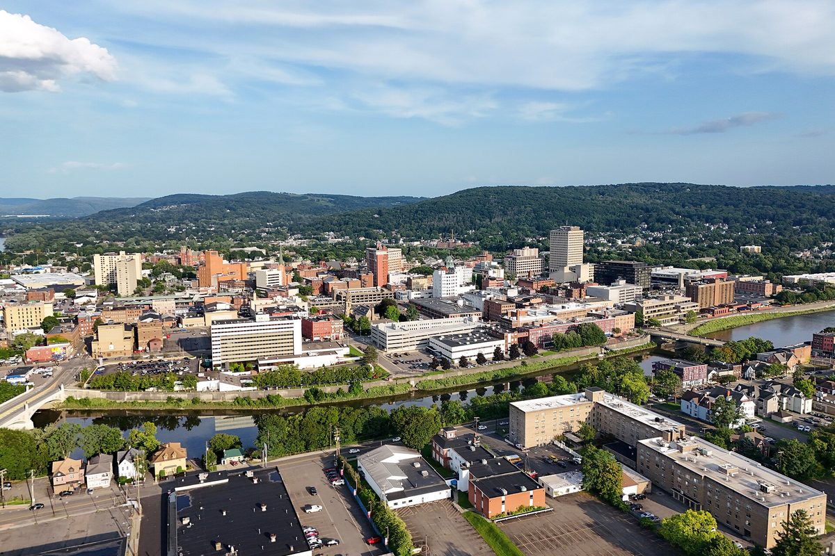 Aerial view of downtown Binghamton beside the river, with wooded hills rising behind the city under a partly cloudy sky