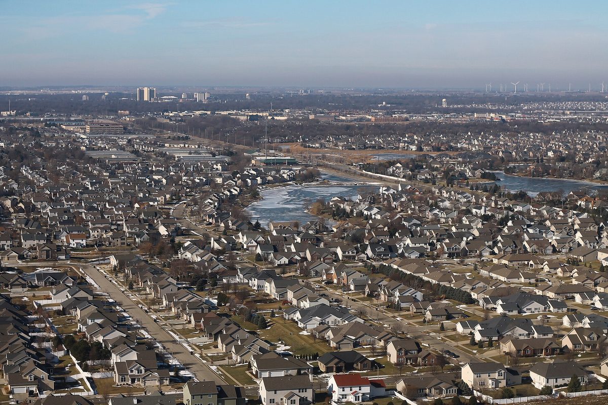 Aerial view over rows of Bloomington houses and a partly frozen lake, with taller buildings and wind turbines on the far horizon