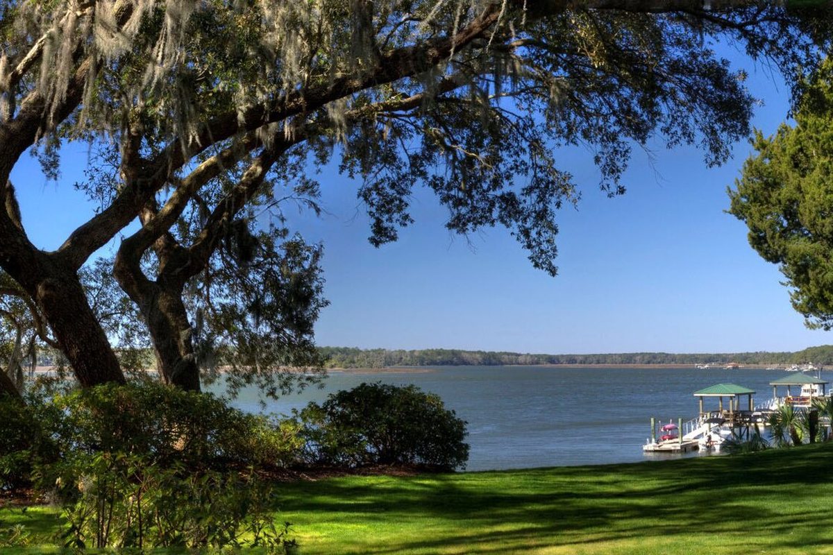 A live oak hung with Spanish moss framing a wide river view in Bluffton, with a green lawn and a covered dock on the water