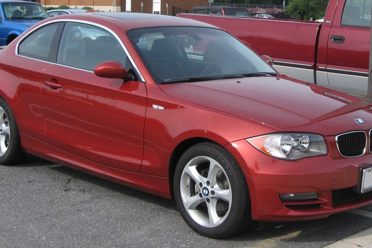 A red 2008 BMW 128i coupe with silver five-spoke wheels, parked in a lot beside a red pickup truck