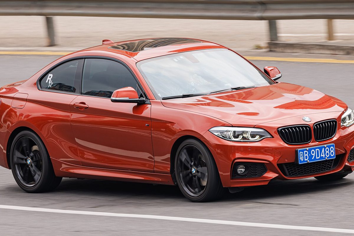An orange first-generation BMW 2 Series coupe with black wheels, driving past a highway guardrail