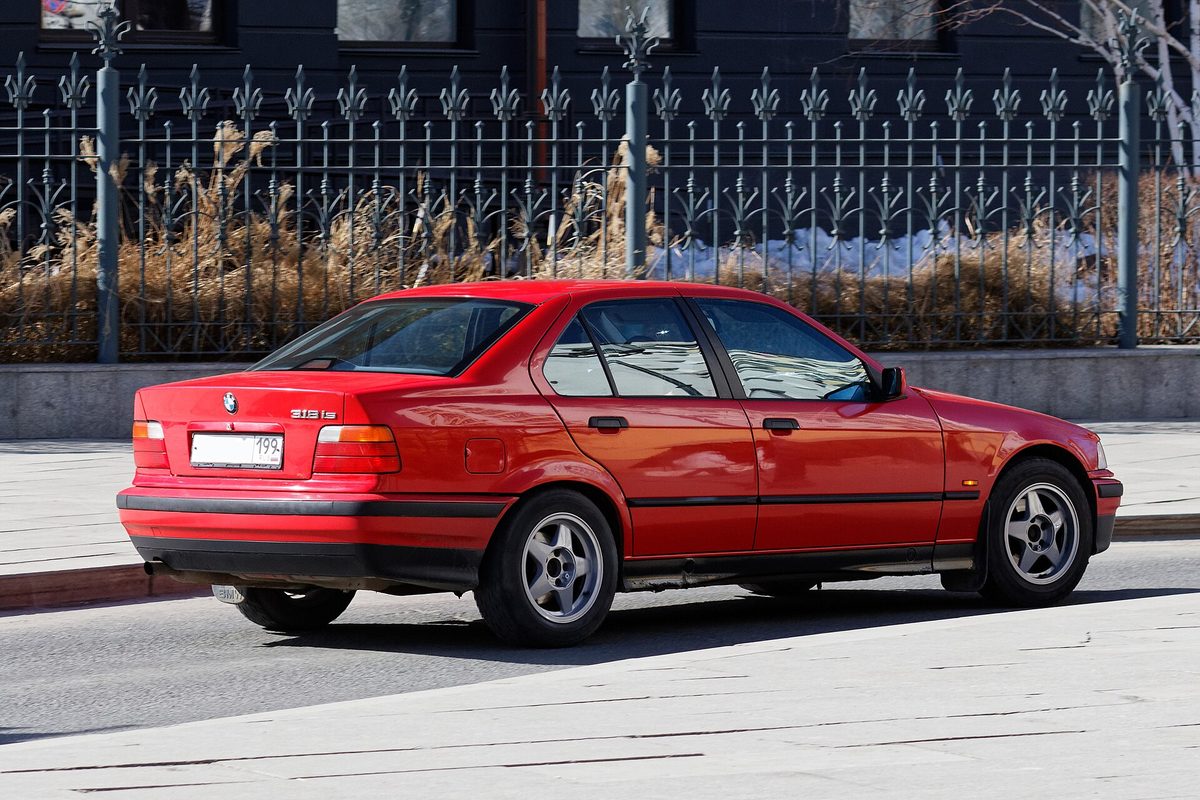 A red 1990s BMW 3 Series sedan seen from the rear quarter, parked on a city street beside a black iron fence