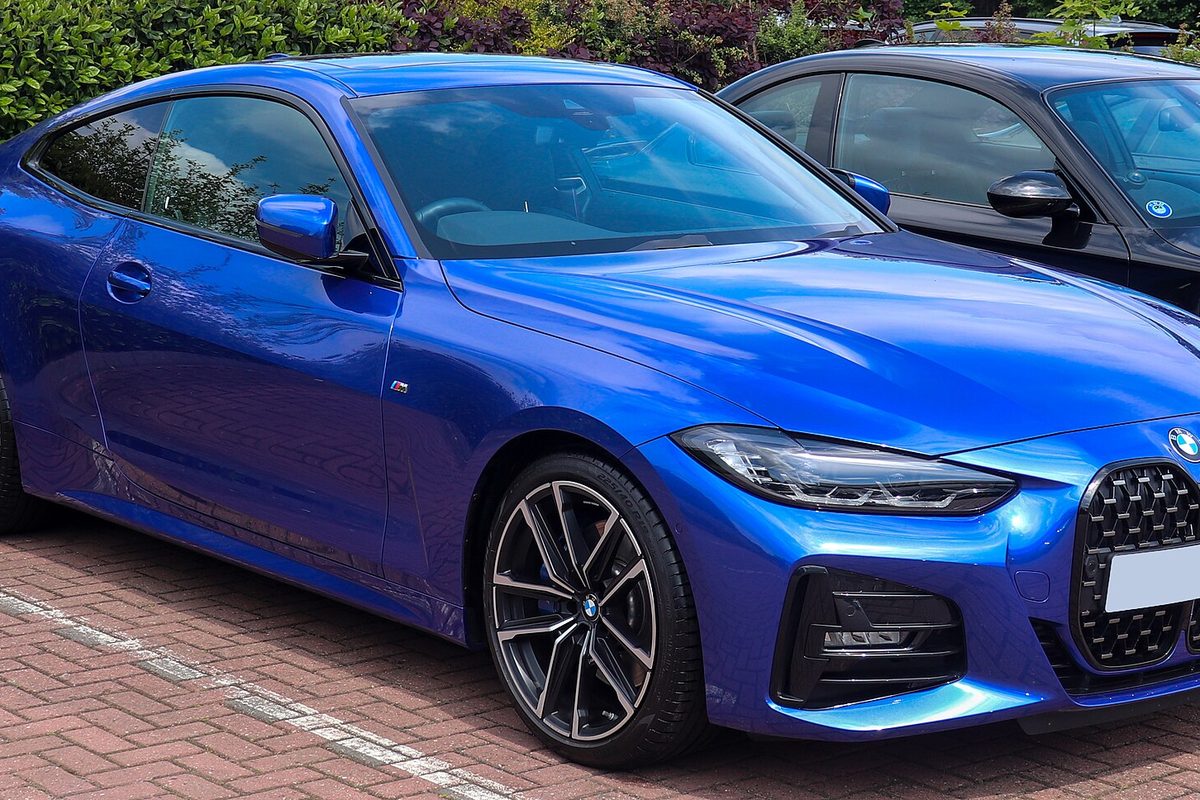 A bright blue second-generation BMW 4 Series coupe with a tall kidney grille and dark multi-spoke wheels, parked on brick paving by a hedge