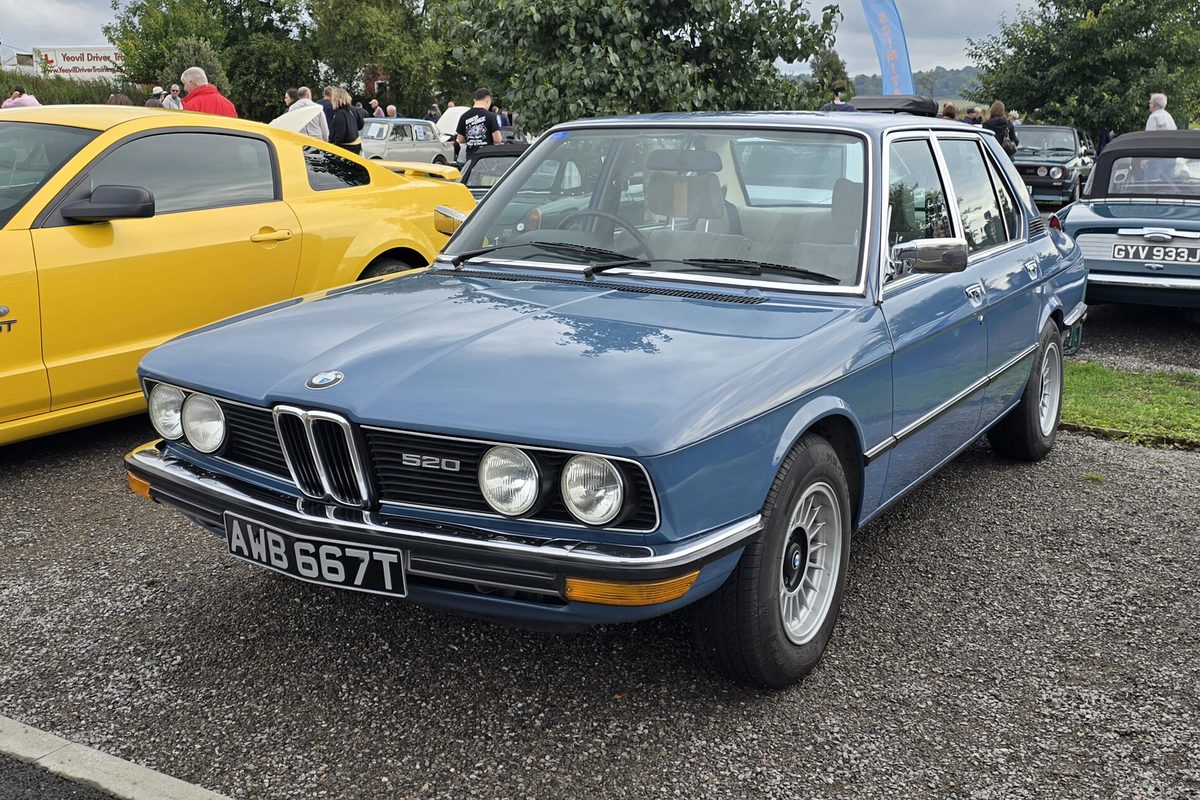 A blue 1970s BMW 520 sedan with four round headlights and chrome bumpers, parked on gravel at a car show