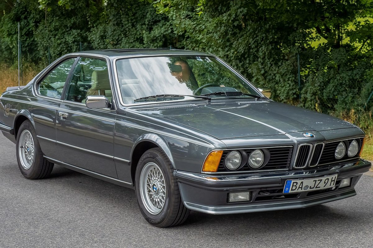 A dark gray first-generation BMW 635CSi coupe with cross-spoke alloy wheels on a tree-lined road