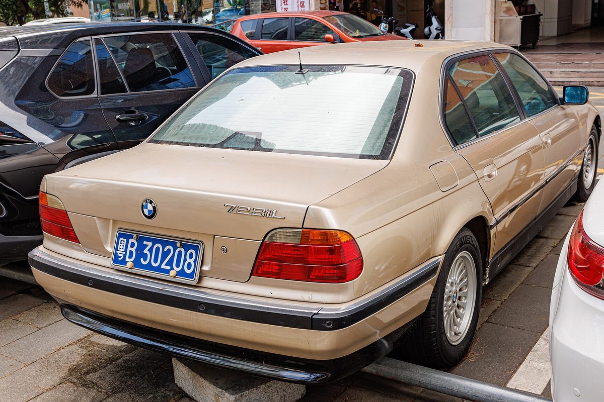 A champagne-gold BMW 728iL long-wheelbase sedan from the E38 generation, seen from the rear, parked with one wheel on a curb