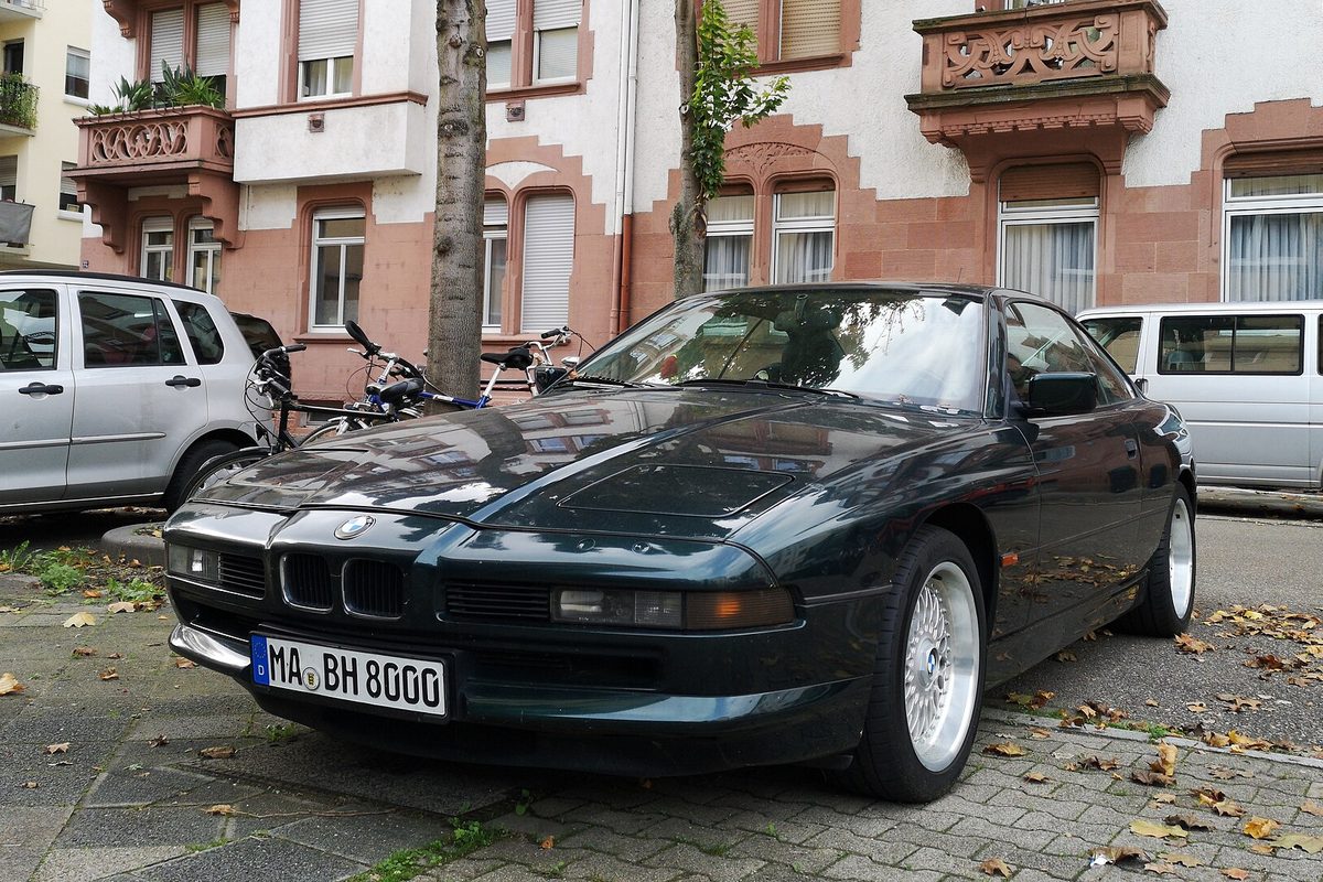 A dark green first-generation BMW 8 Series coupe with silver mesh wheels parked on a cobbled sidewalk with fallen leaves