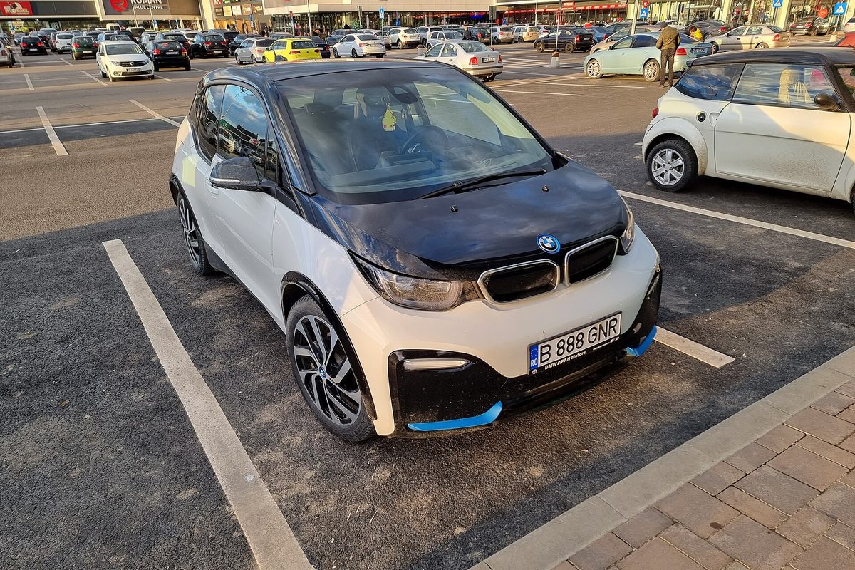 A white BMW i3 hatchback with a black hood and blue bumper trim, parked in a shopping center lot