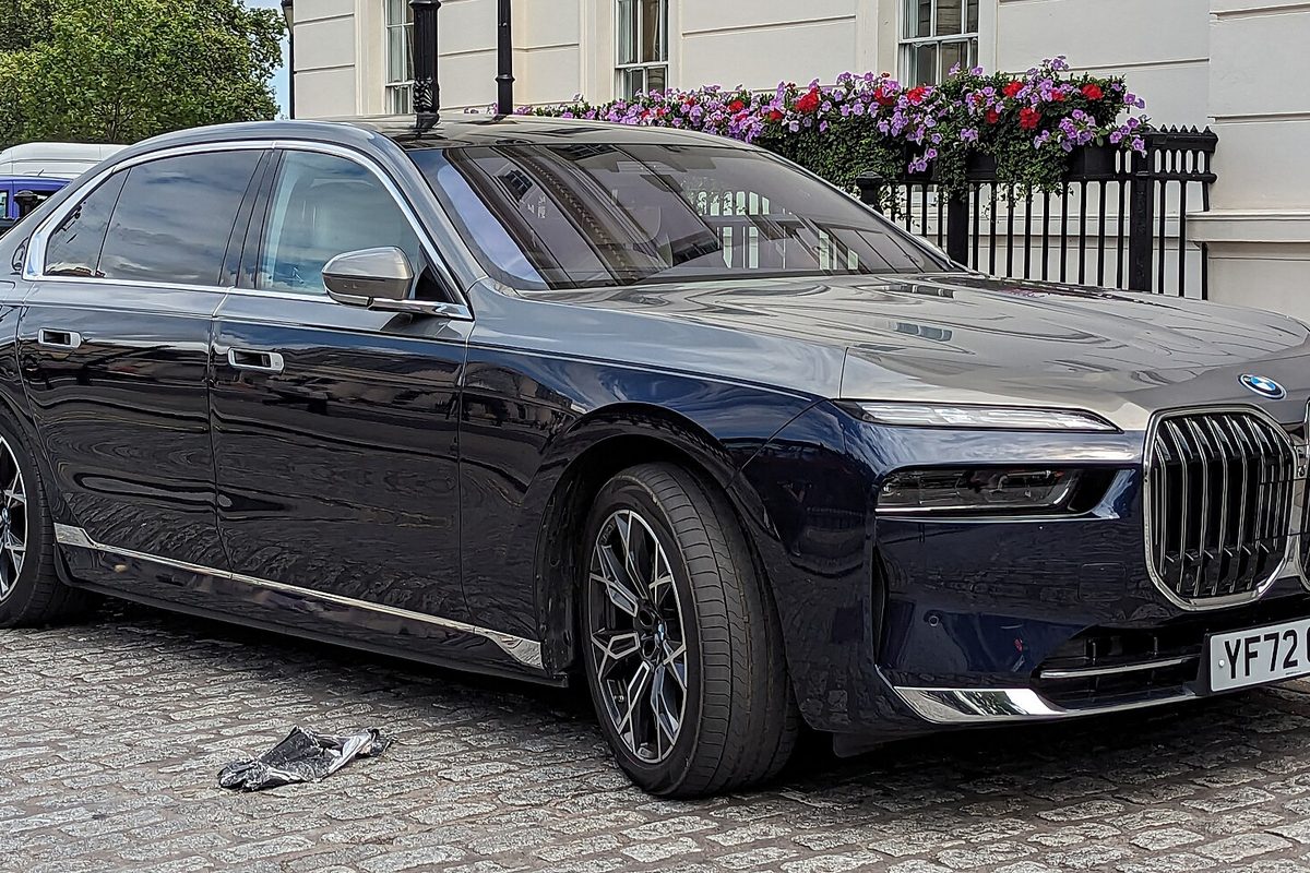 A dark blue BMW i7 sedan with a tall grille parked on cobblestones beside a railing hung with flower boxes