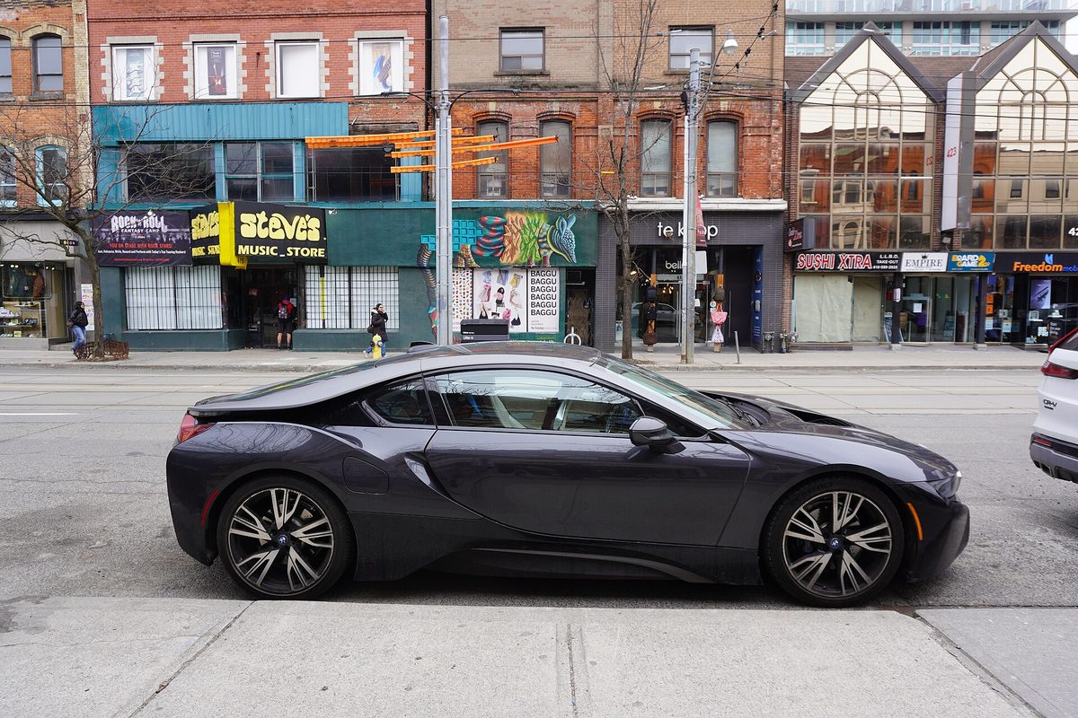 A dark gray BMW i8 coupe parked at a city curb in front of a row of shops