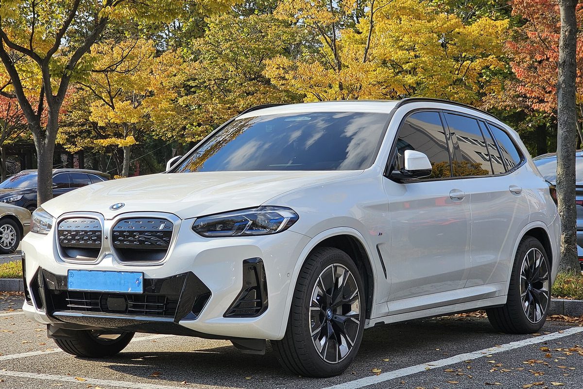 A white earlier-generation (G08) BMW iX3 electric SUV parked under yellow autumn trees
