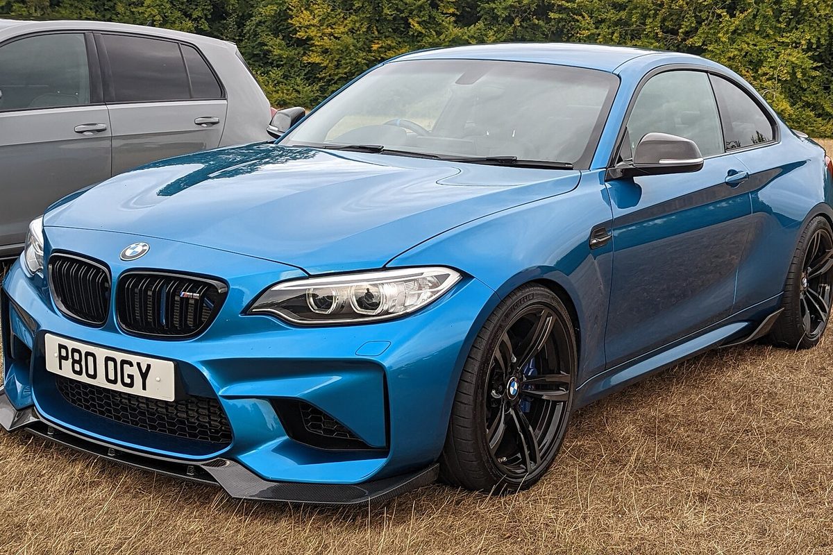 A bright blue first-generation BMW M2 coupe with black wheels and a front splitter parked on dry grass