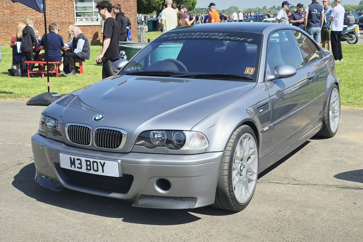 A gray E46-generation BMW M3 coupe with silver alloy wheels at an outdoor car show, spectators and tents on the grass behind it
