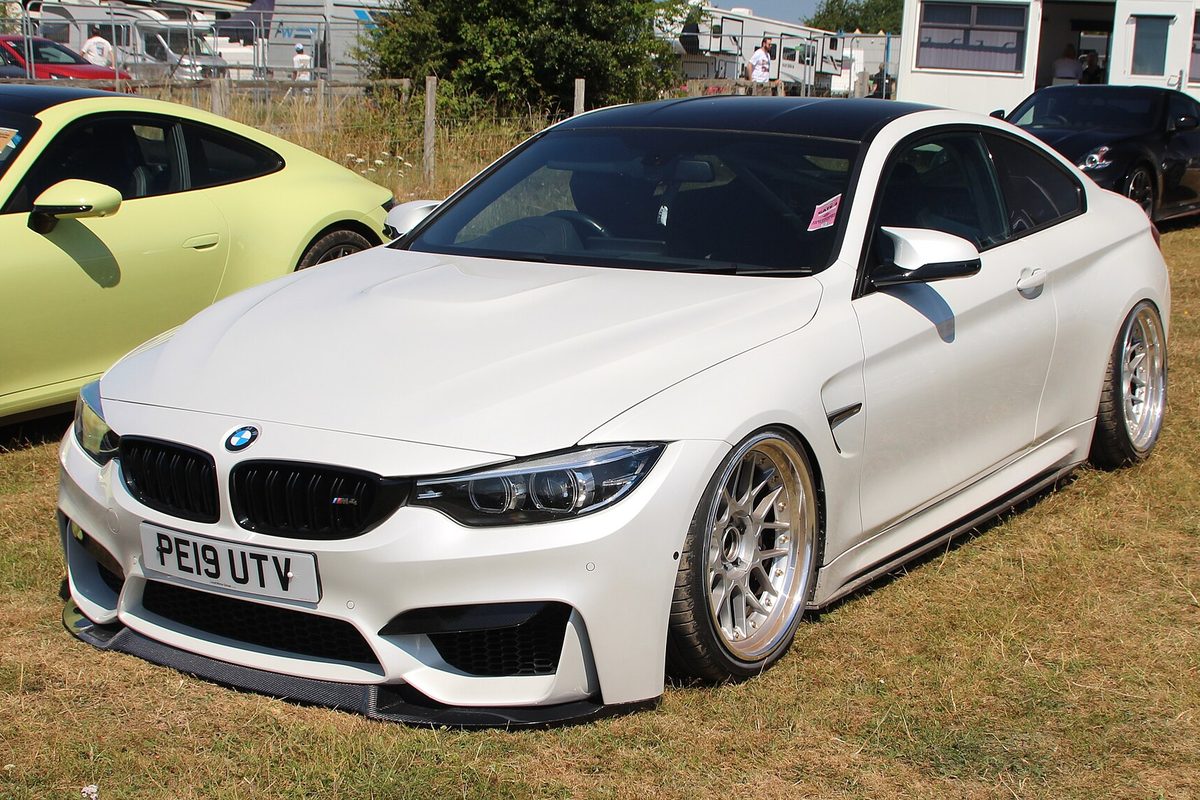 A lowered white first-generation BMW M4 coupe with a black roof and silver mesh wheels parked on dry grass at a car show