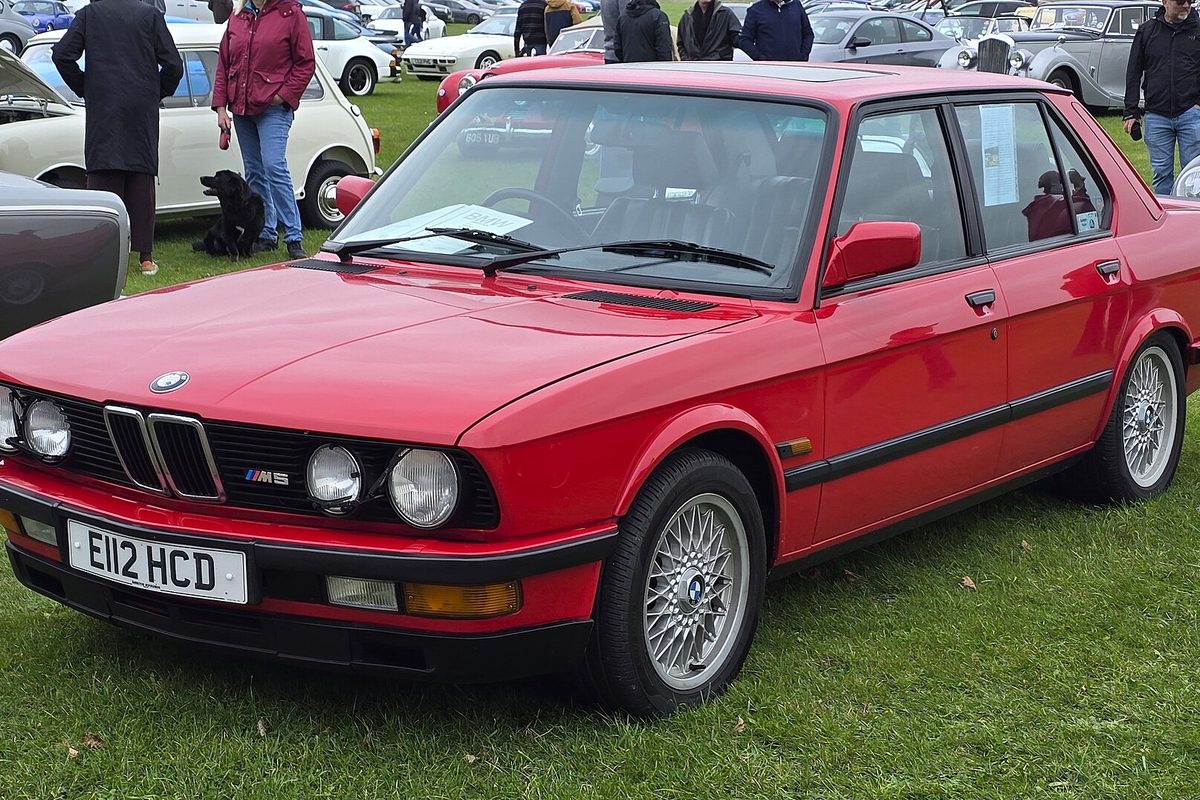 A red first-generation (E28) BMW M5 sedan parked on grass at a car show