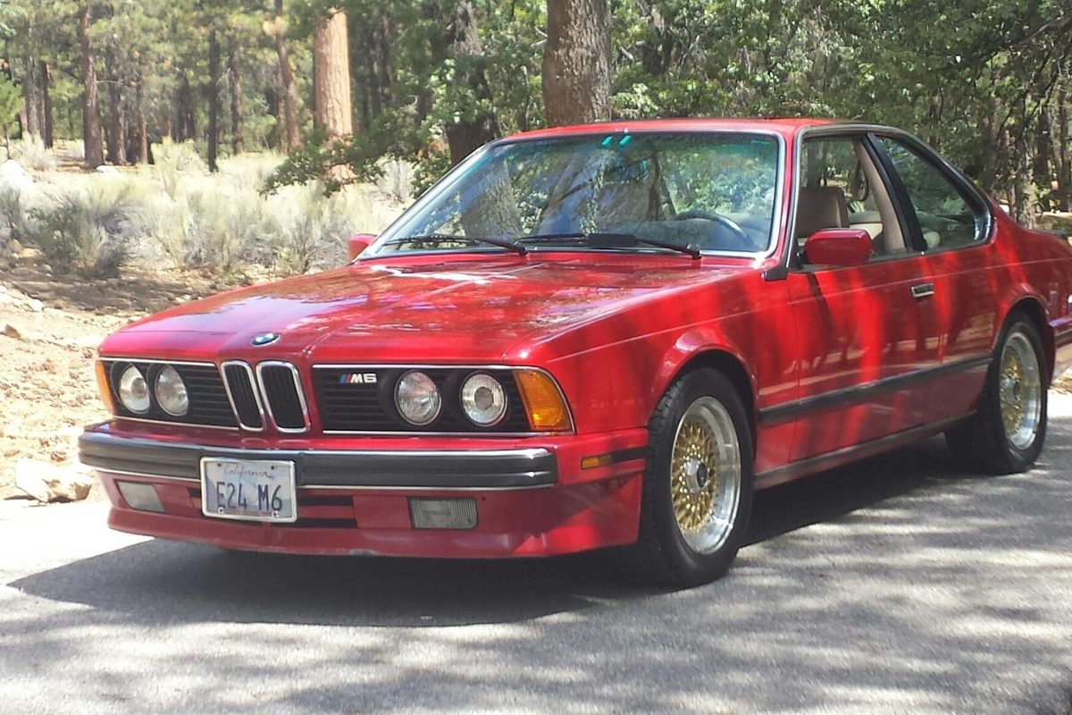 A red 1988 BMW M6 coupe with gold mesh wheels parked on a shaded road among pine trees