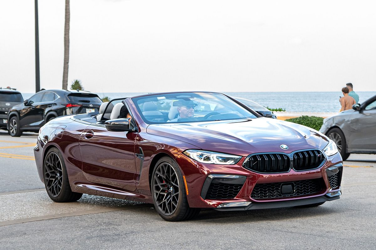 A dark red BMW M8 convertible with the top down and black wheels, driving along a beachfront road