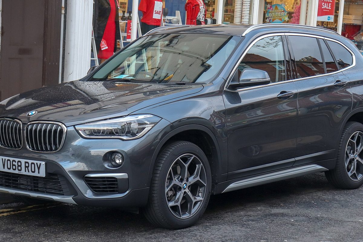 A dark gray second-generation BMW X1 with silver roof rails and two-tone alloy wheels, parked on a wet street in front of shop windows