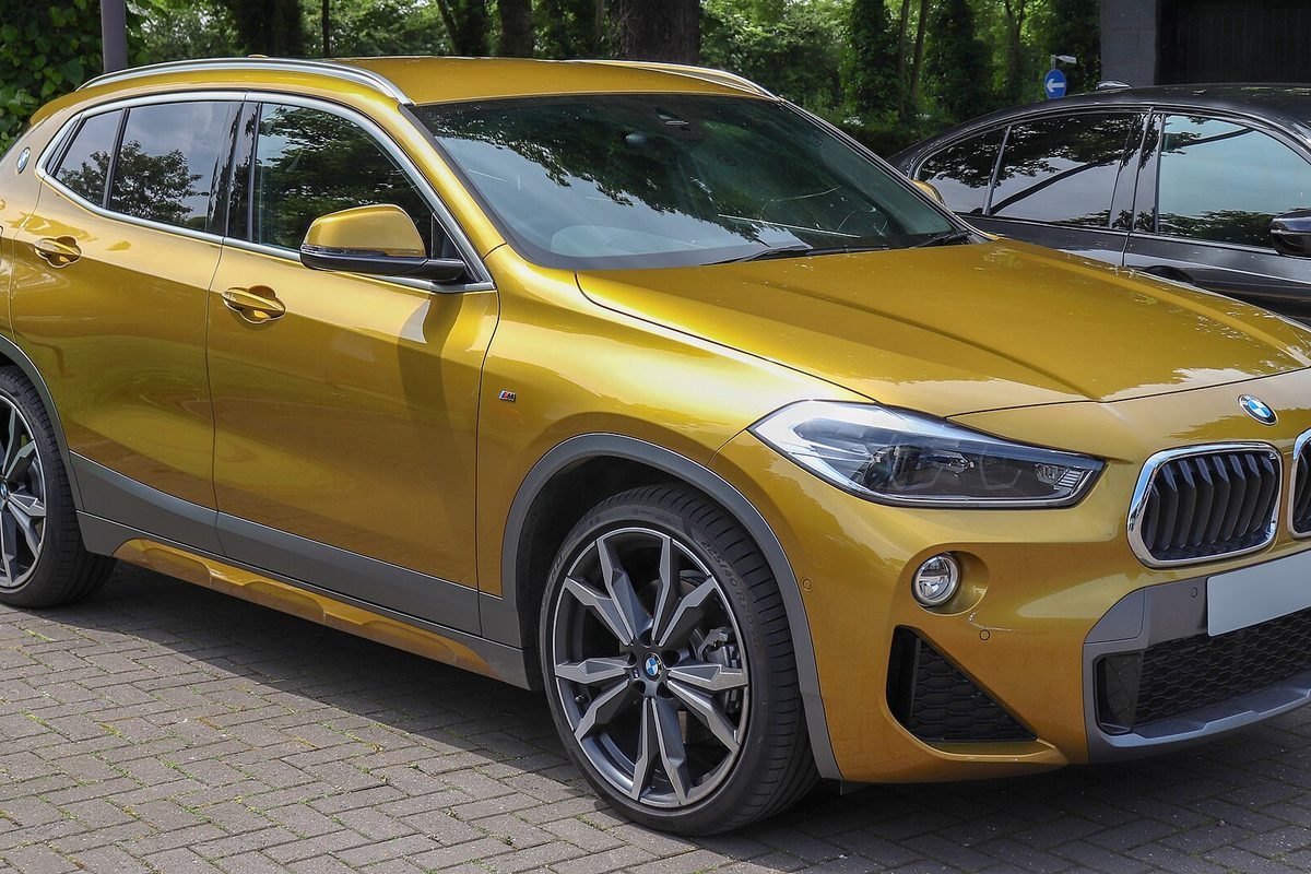 A metallic gold first-generation BMW X2 parked on brick paving with trees behind it, seen from the front right