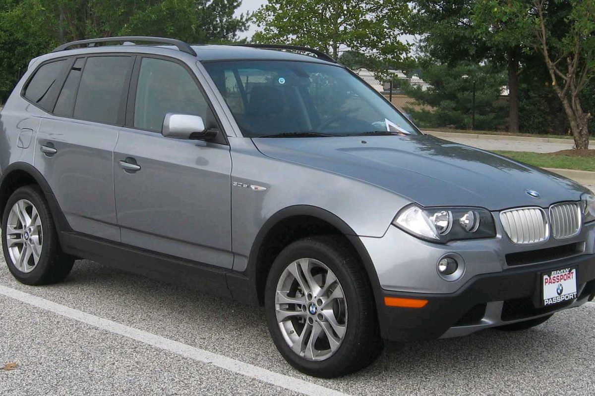 A gray first-generation BMW X3 compact SUV with roof rails, front three-quarter view, parked in a lot with trees behind it
