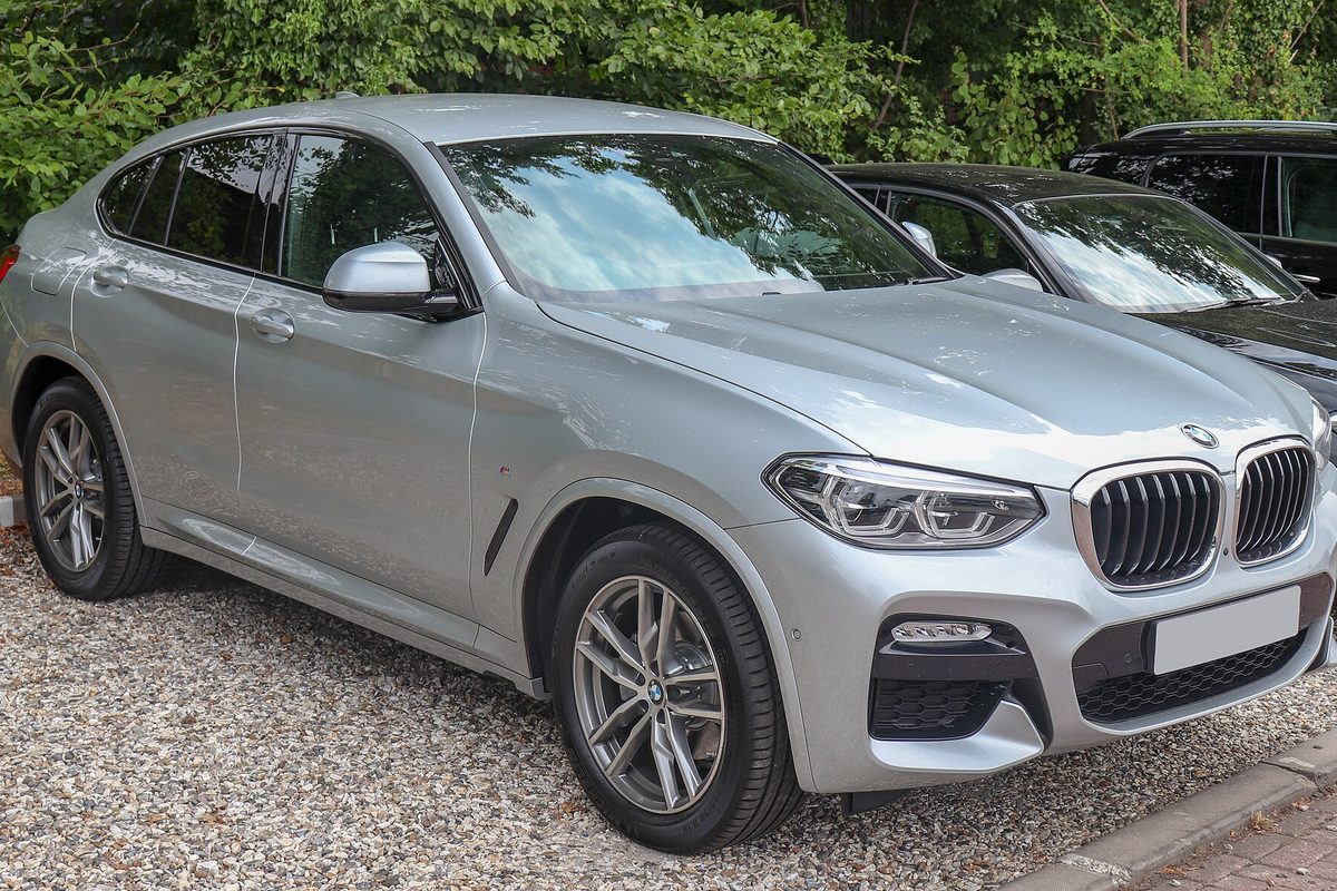 A silver BMW X4 with M Sport bumpers parked on gravel in front of green bushes