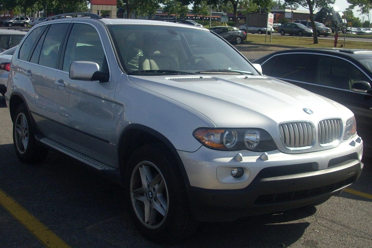 A silver first-generation BMW X5 with a silver grille, parked in a sunny lot with trees and buildings behind it