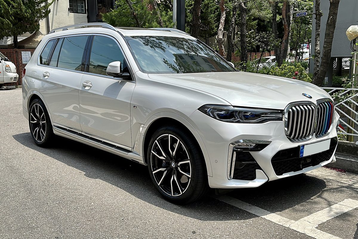 A white BMW X7 SUV with a large chrome kidney grille and black two-tone wheels, parked in a lot under trees