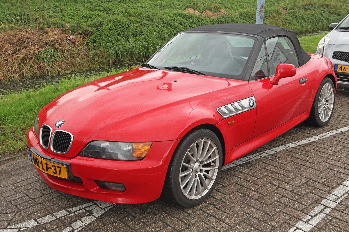 A red 1996 BMW Z3 roadster with its black soft top up and silver multi-spoke wheels, parked on paving stones beside a grassy bank