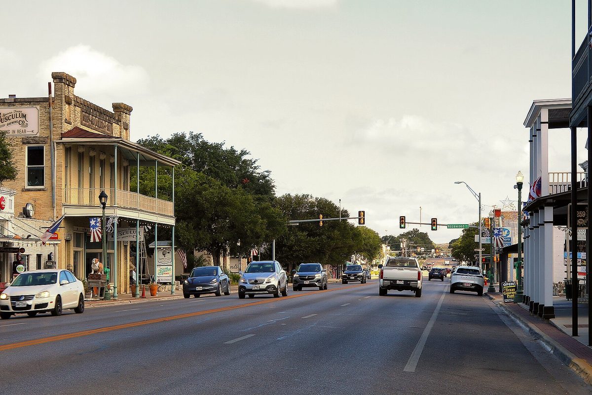 Main Street in Boerne on a summer afternoon, with cars passing two-story stone and wood storefronts with balconies, flags and black lamp posts