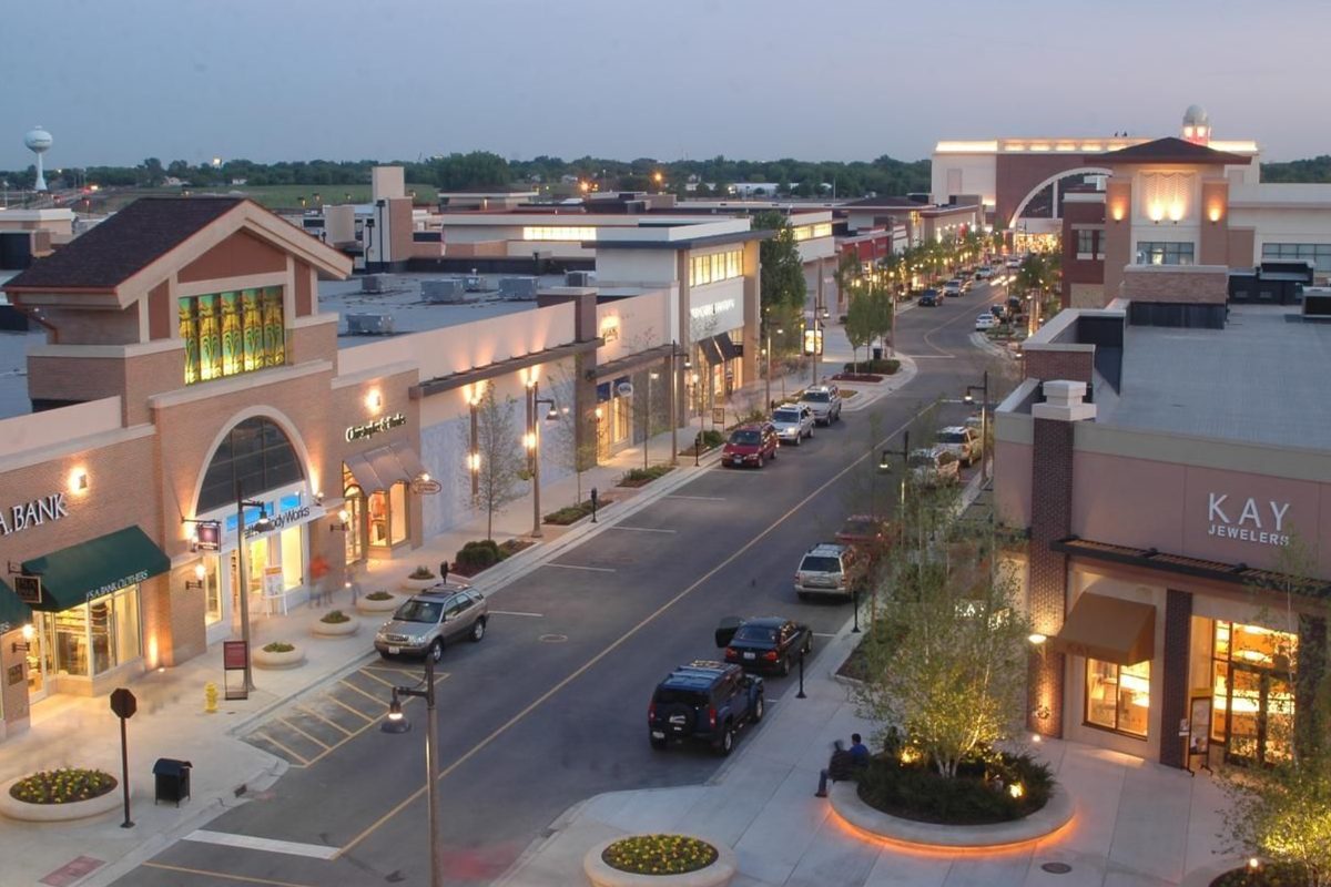 An outdoor shopping street in Bolingbrook at dusk, with lit storefronts on both sides, cars on the road down the middle and a water tower on the skyline