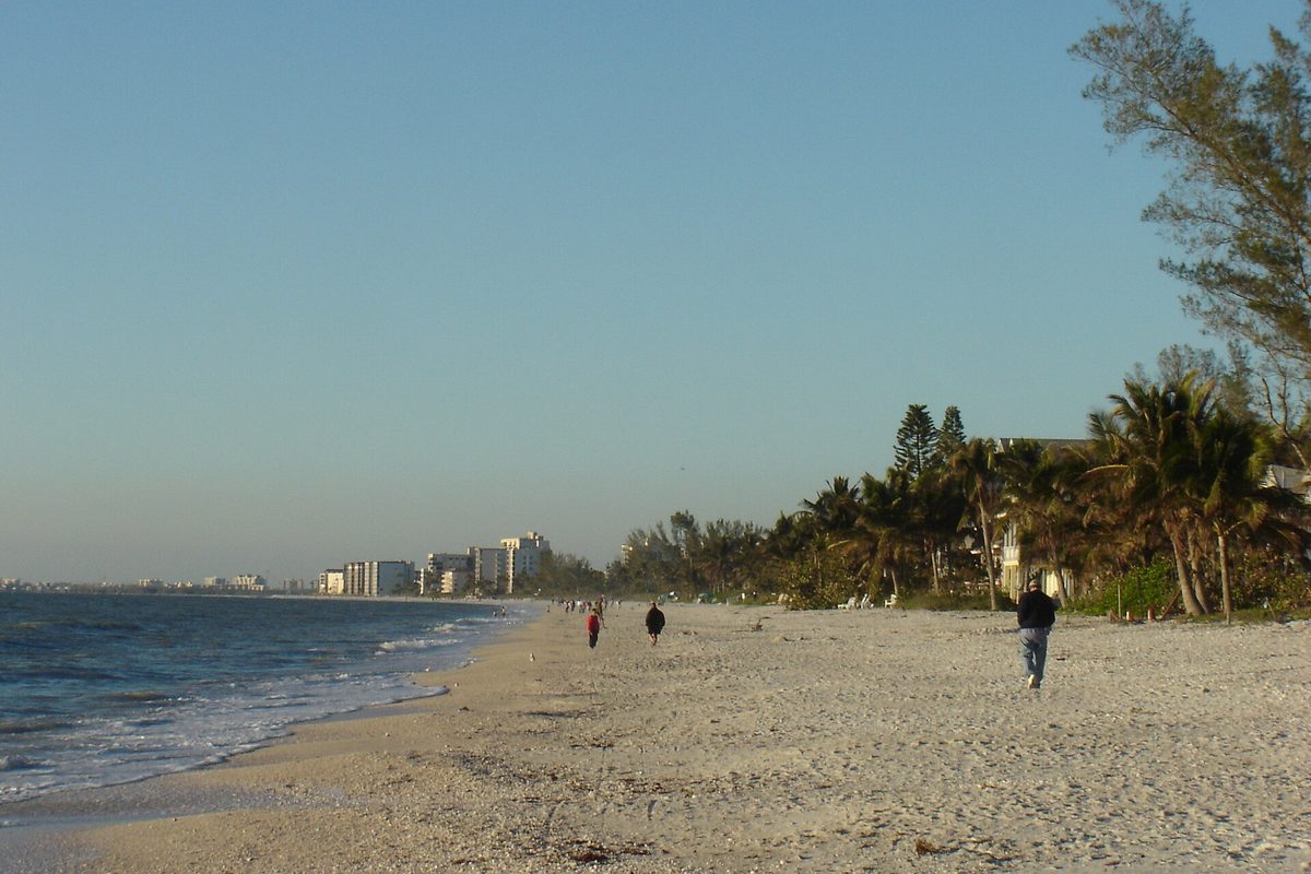 People walking on the wide white sand of Bonita Beach at the water's edge, with palm trees on the right and high-rise buildings in the distance